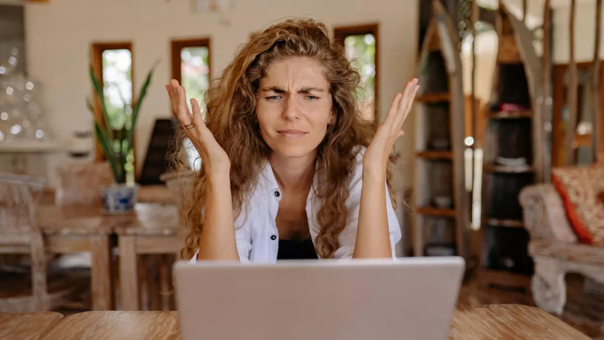 A woman sits at a wooden table, looking frustrated as she works on a laptop. Her arms are raised in a gesture of exasperation. A smartphone lies on the table beside the laptop.