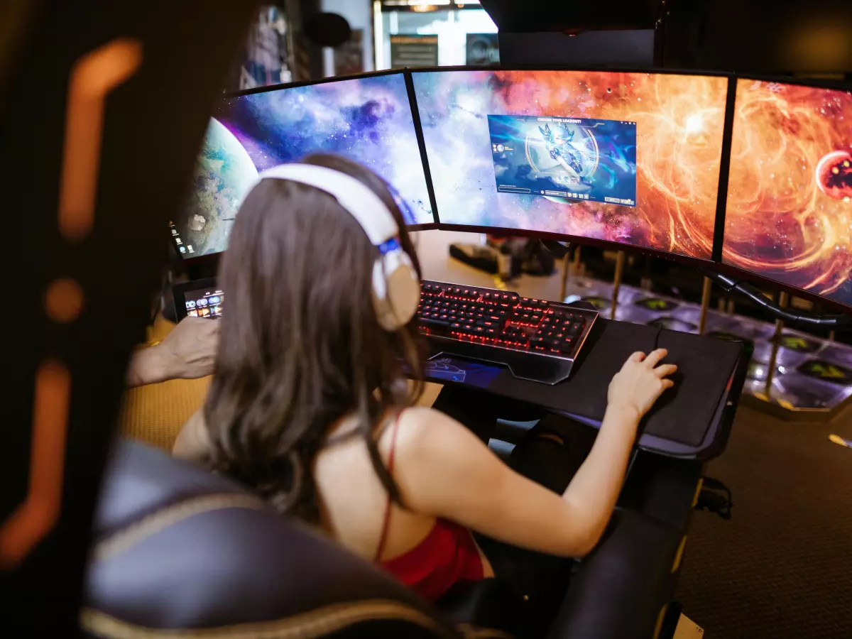 A woman wearing a headset plays a video game on a triple monitor setup.