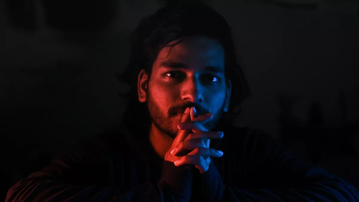 A young man sitting at a table, illuminated in blue and red light, holding a video game controller. He appears to be focused on the game. The image captures the immersive nature of gaming and the use of dynamic lighting effects.