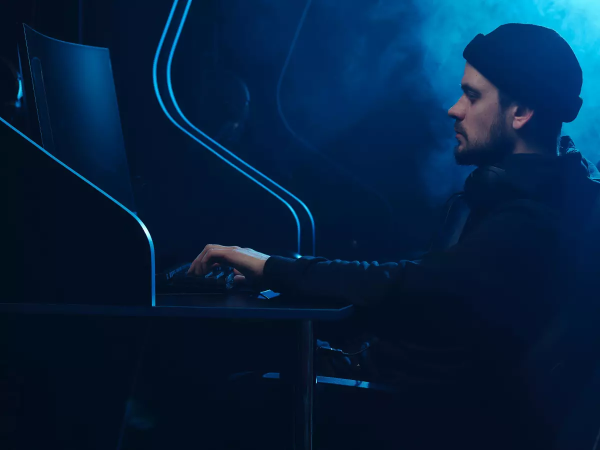 A young man with a beard and black beanie is sitting in a dark room and typing on a computer keyboard. The room is lit with a blue and white glow.