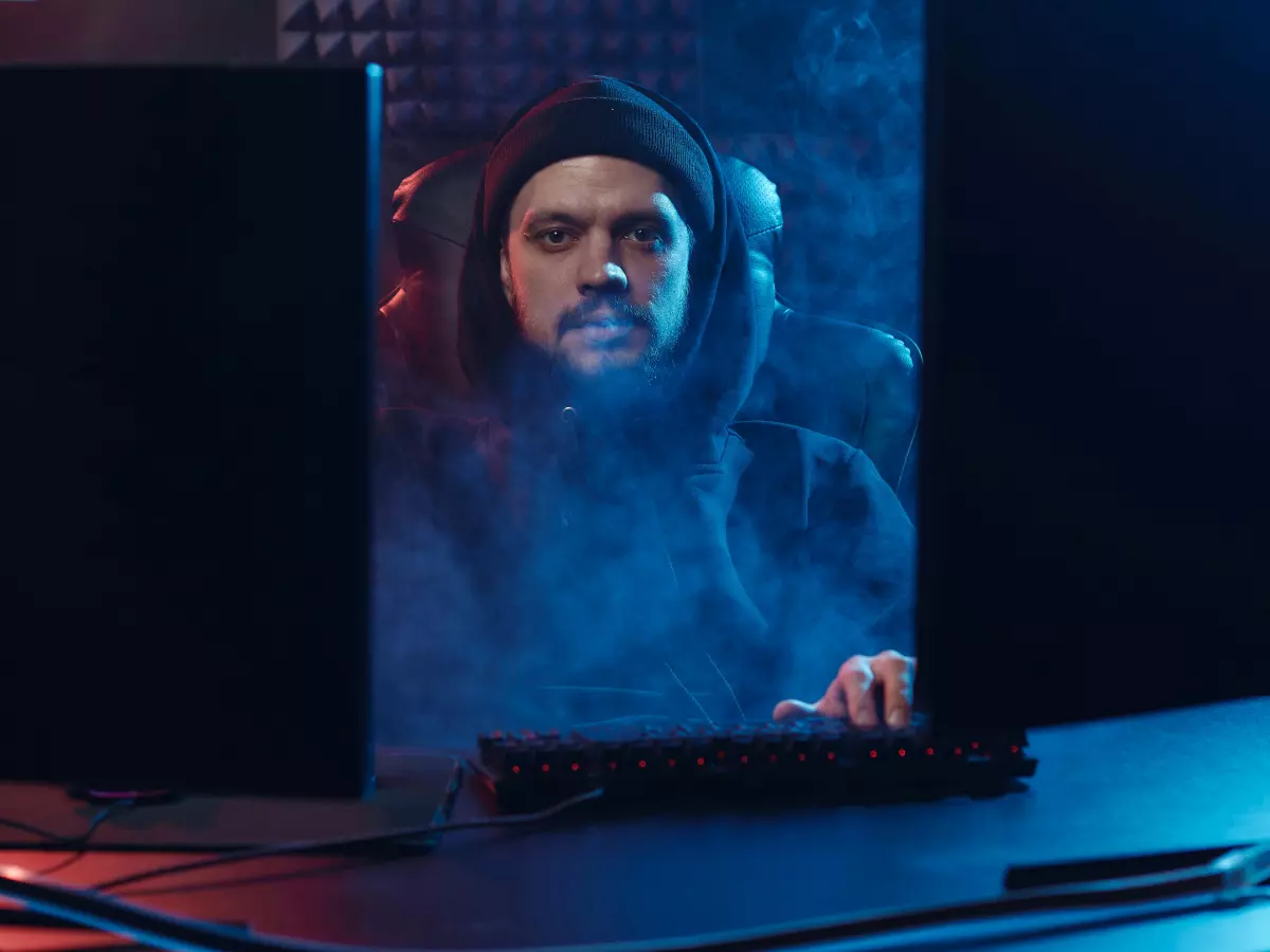 A hooded figure sits in front of a computer in a dimly lit room, typing with a determined look.  A blue glow illuminates the scene and there is a feeling of mystery and danger.