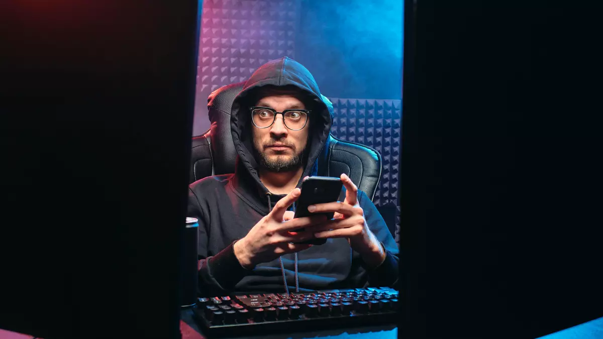 A person in a dark hoodie sits in front of two computer monitors, holding a smartphone in their hand. The room is lit with blue and red lights, creating a dramatic atmosphere.