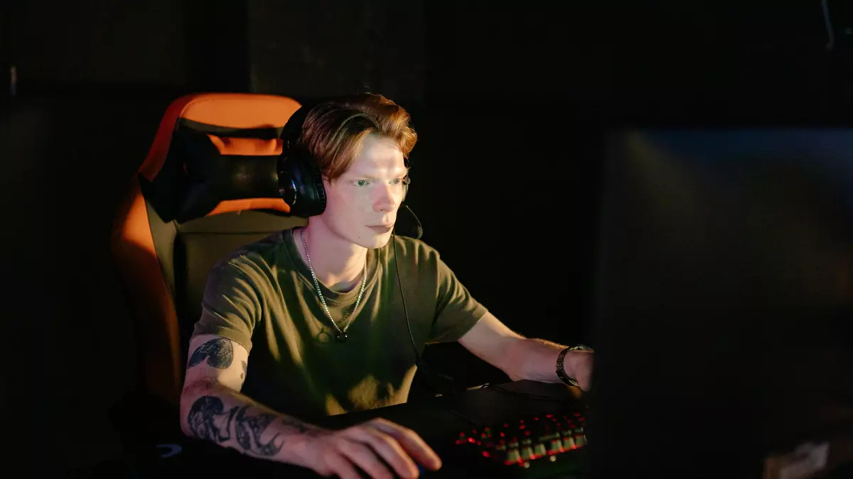 A young man wearing a headset is seated in front of a computer, focused on gaming. He