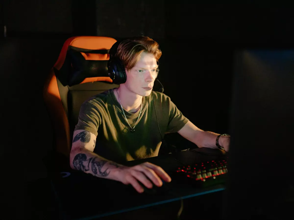 A young man wearing a headset is seated in front of a computer, focused on gaming. He's illuminated by the screen light in a darkened room.