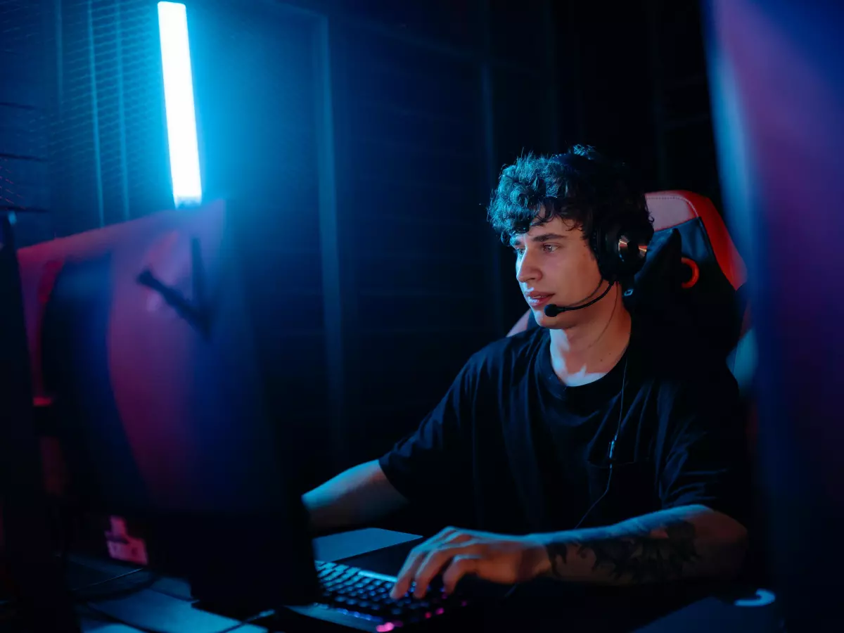 A man is sitting in a dark room with a headset on, using a computer. The room is lit up with blue and purple lights.