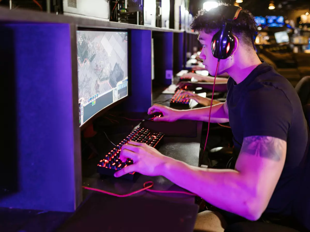 A young man is sitting in front of a computer wearing a headset, concentrating on the screen. The room is lit with a purple hue, enhancing the sense of immersion in the gaming world. The image is dramatic and visually appealing.