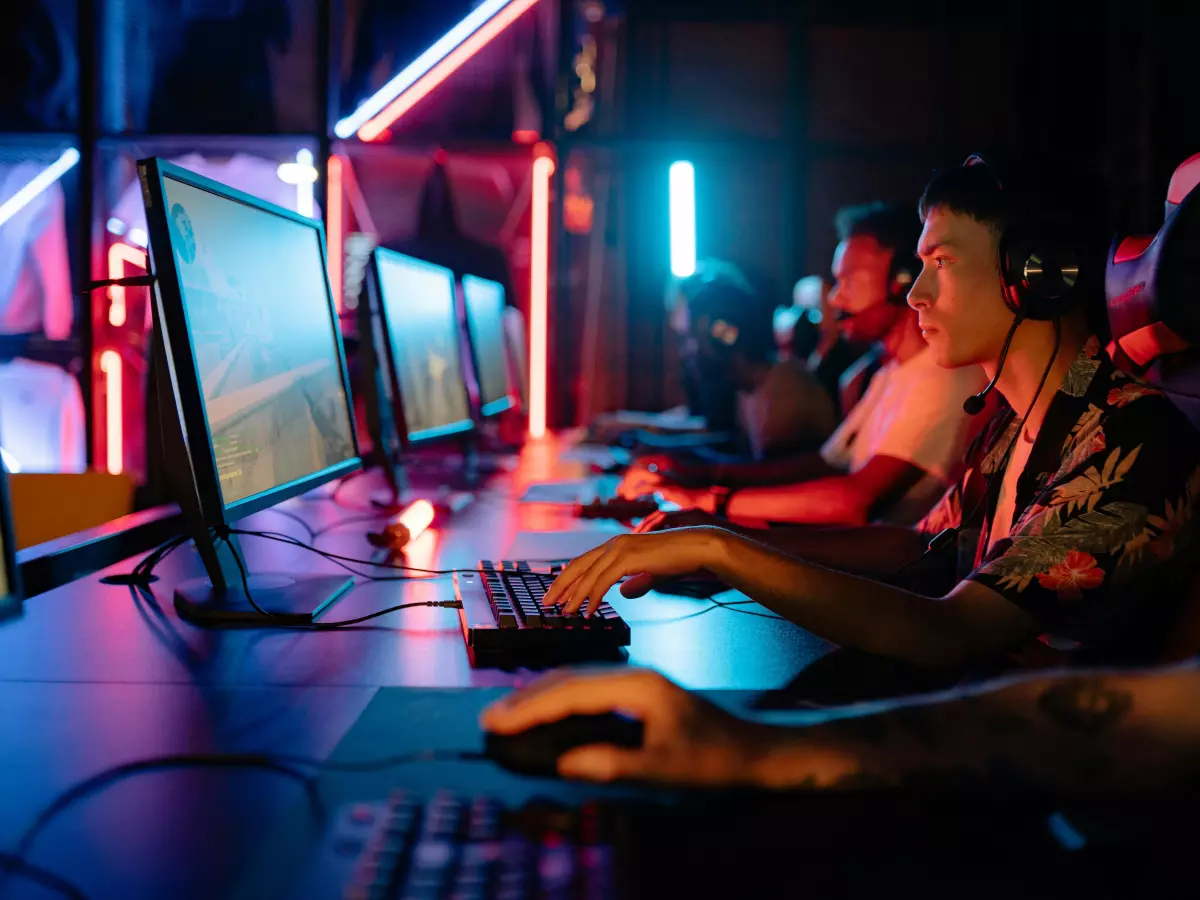 A group of young men are playing video games in a dark room with neon lights. They are all focused on their screens and have headsets on.