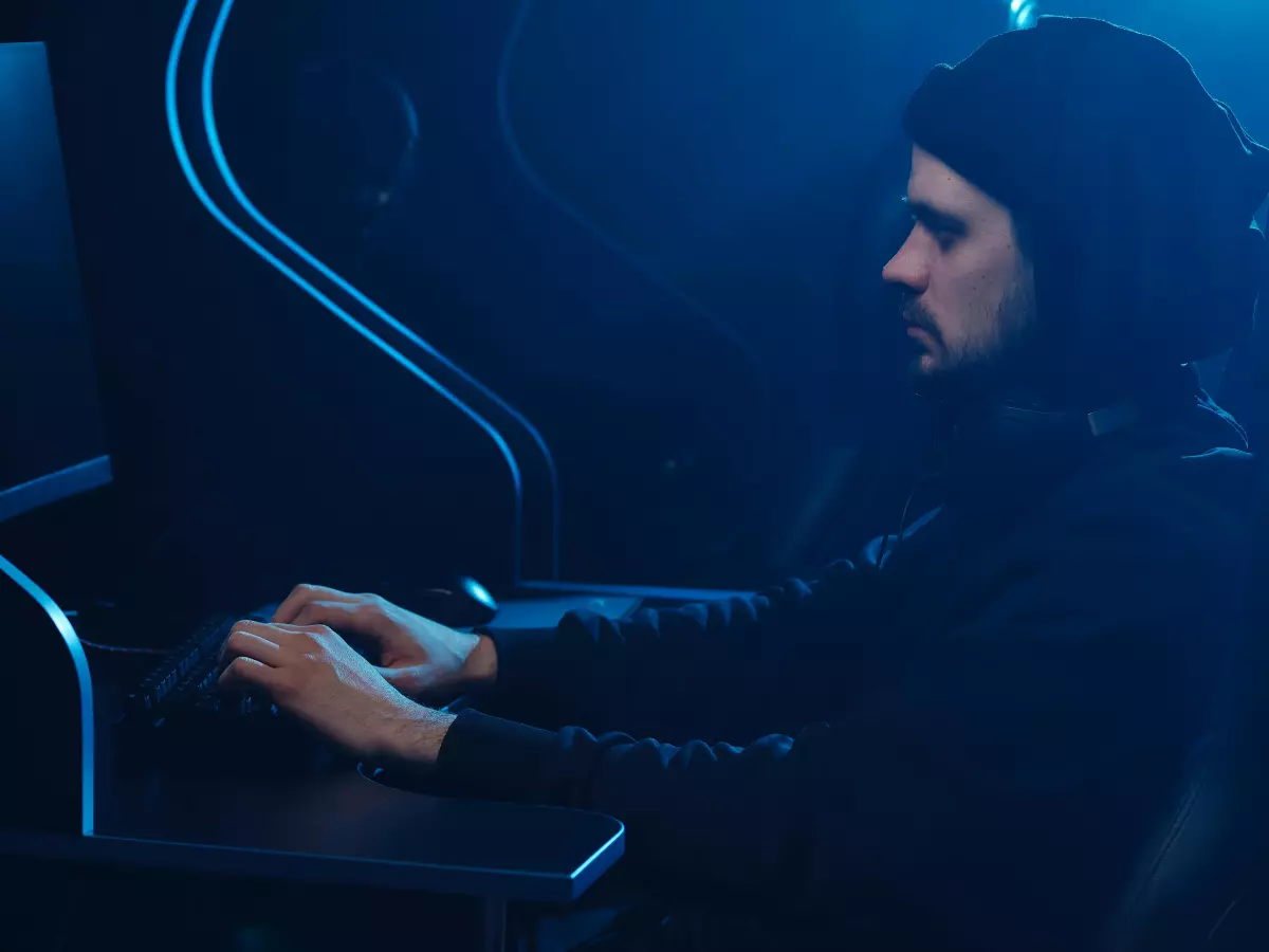 A hooded figure sits in a dimly lit room, typing on a computer keyboard. The room is bathed in blue light, casting long shadows.