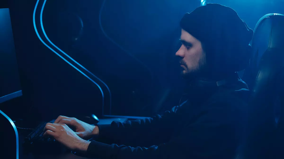 A hooded figure sits in a dimly lit room, typing on a computer keyboard. The room is bathed in blue light, casting long shadows.