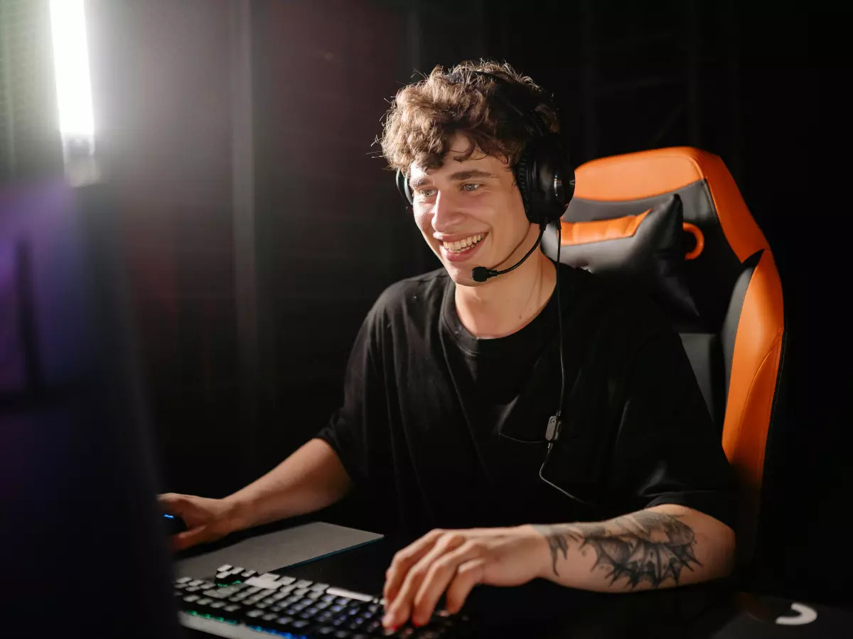 A young man with a headset on, sitting in front of a computer, playing a game. He is smiling and looking at the screen.