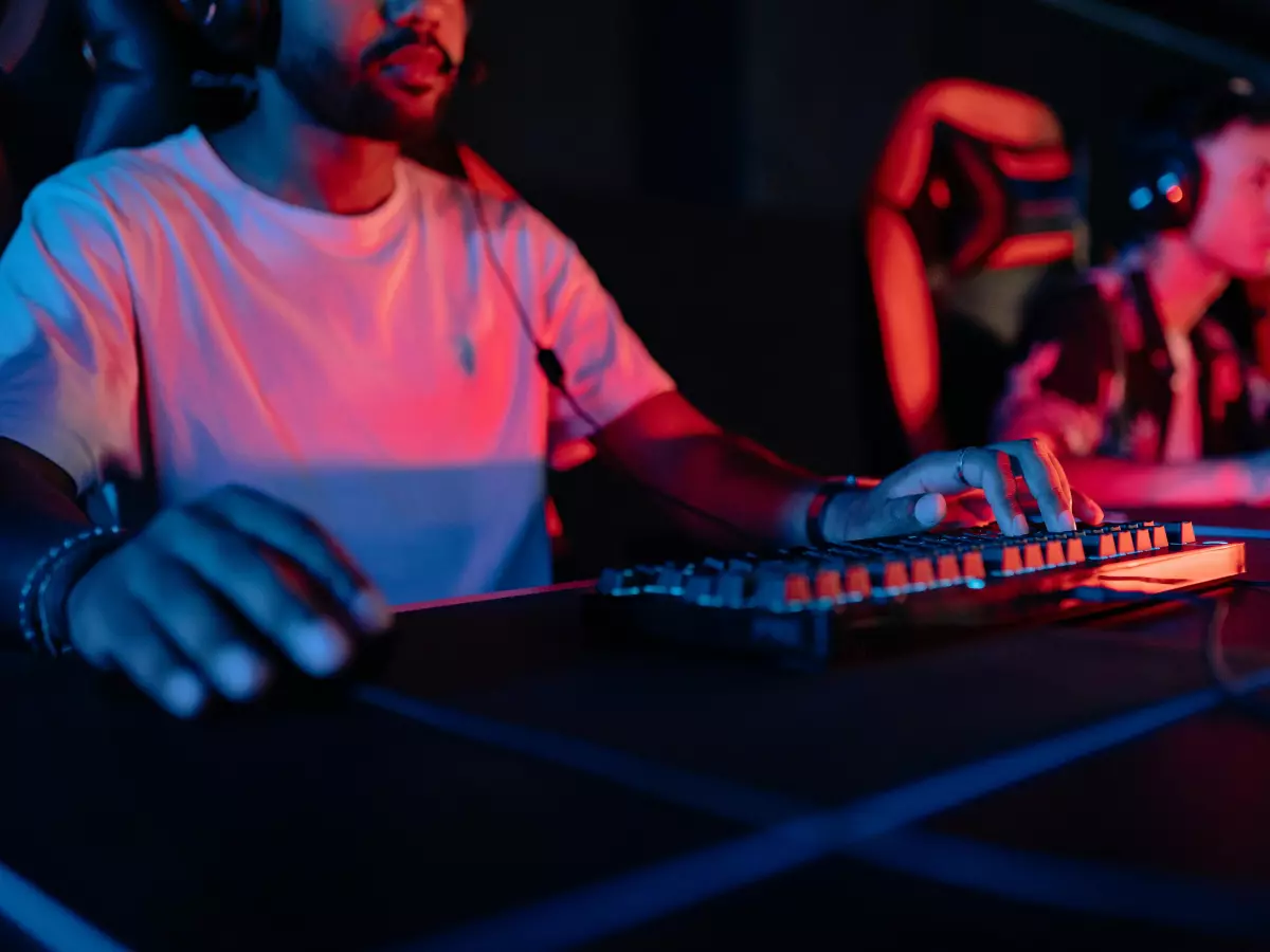 Close up of a person's hand typing on a gaming keyboard. The keyboard is illuminated in red and blue, and the person is wearing a headset.