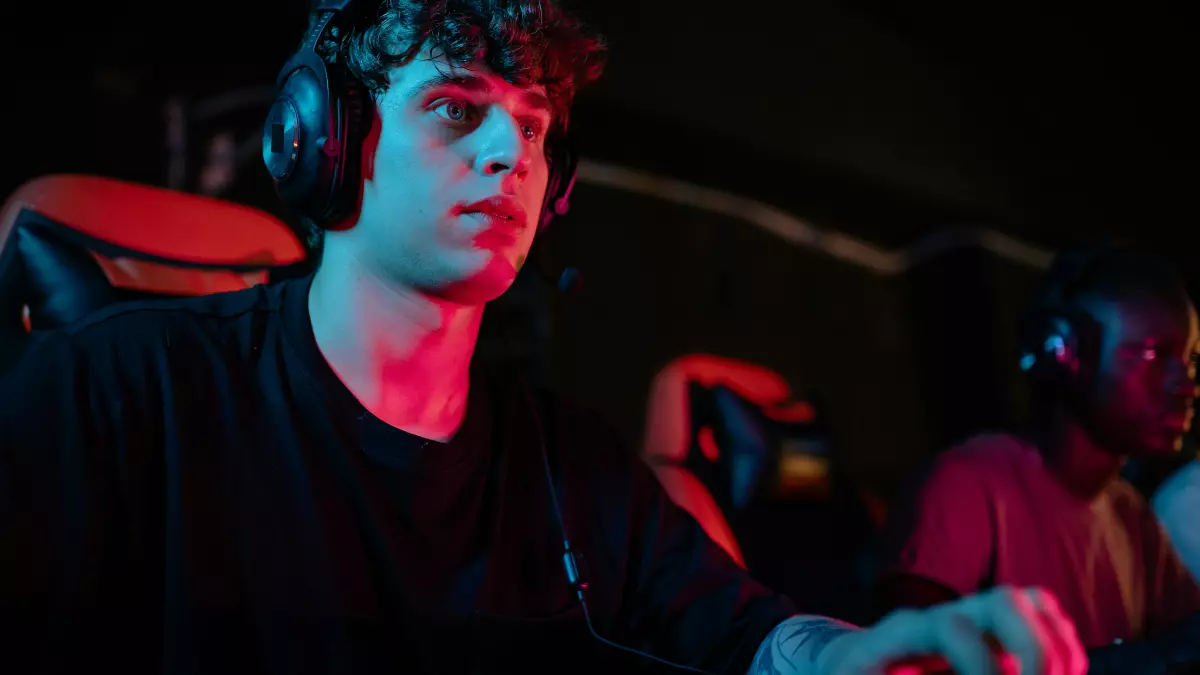 A young man wearing headphones is intently playing a video game on a console with a keyboard in front of him. The room is dark, and the light from the console and the keyboard illuminates his face and hands. He is concentrating hard on the game, and his eyes are focused on the screen. 