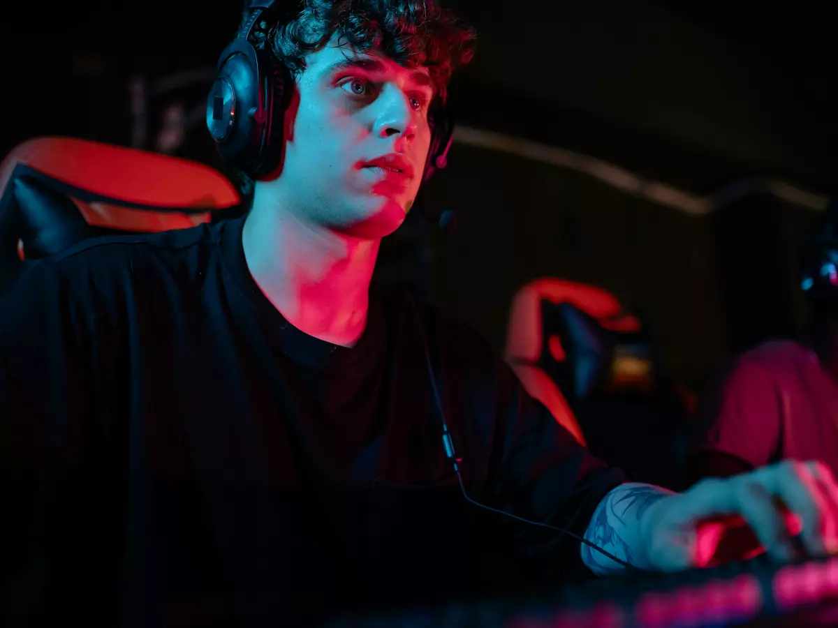 A young man wearing headphones is intently playing a video game on a console with a keyboard in front of him. The room is dark, and the light from the console and the keyboard illuminates his face and hands. He is concentrating hard on the game, and his eyes are focused on the screen. 