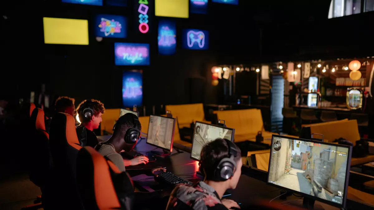 Several people are playing online games in a dark, modern gaming lounge with orange chairs and big monitors, all wearing headphones.