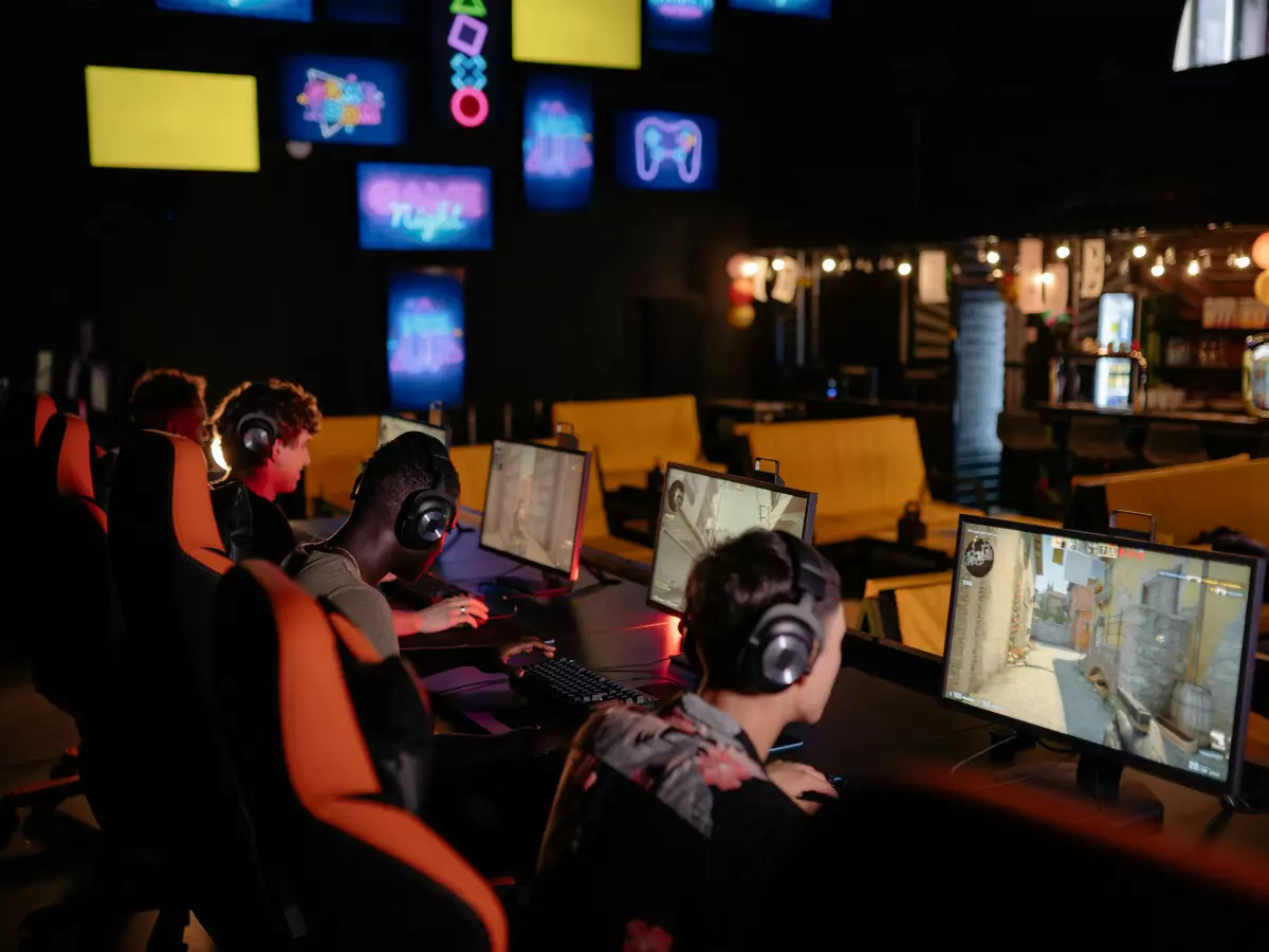 Several people are playing online games in a dark, modern gaming lounge with orange chairs and big monitors, all wearing headphones.