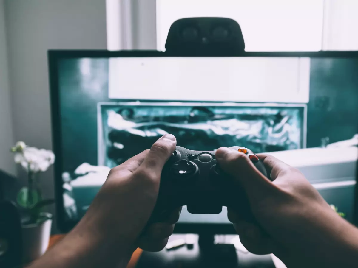 Close-up of hands holding a video game controller, with a monitor showing a game in the background