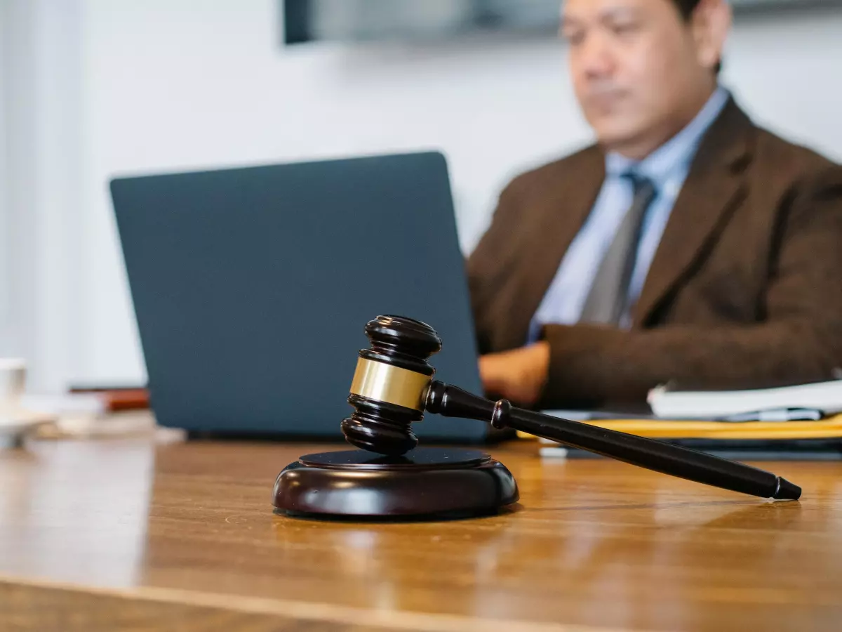 A wooden gavel is placed in the foreground of a desk, with a laptop in the background. A person in a brown suit can be seen in the background, typing on the laptop.