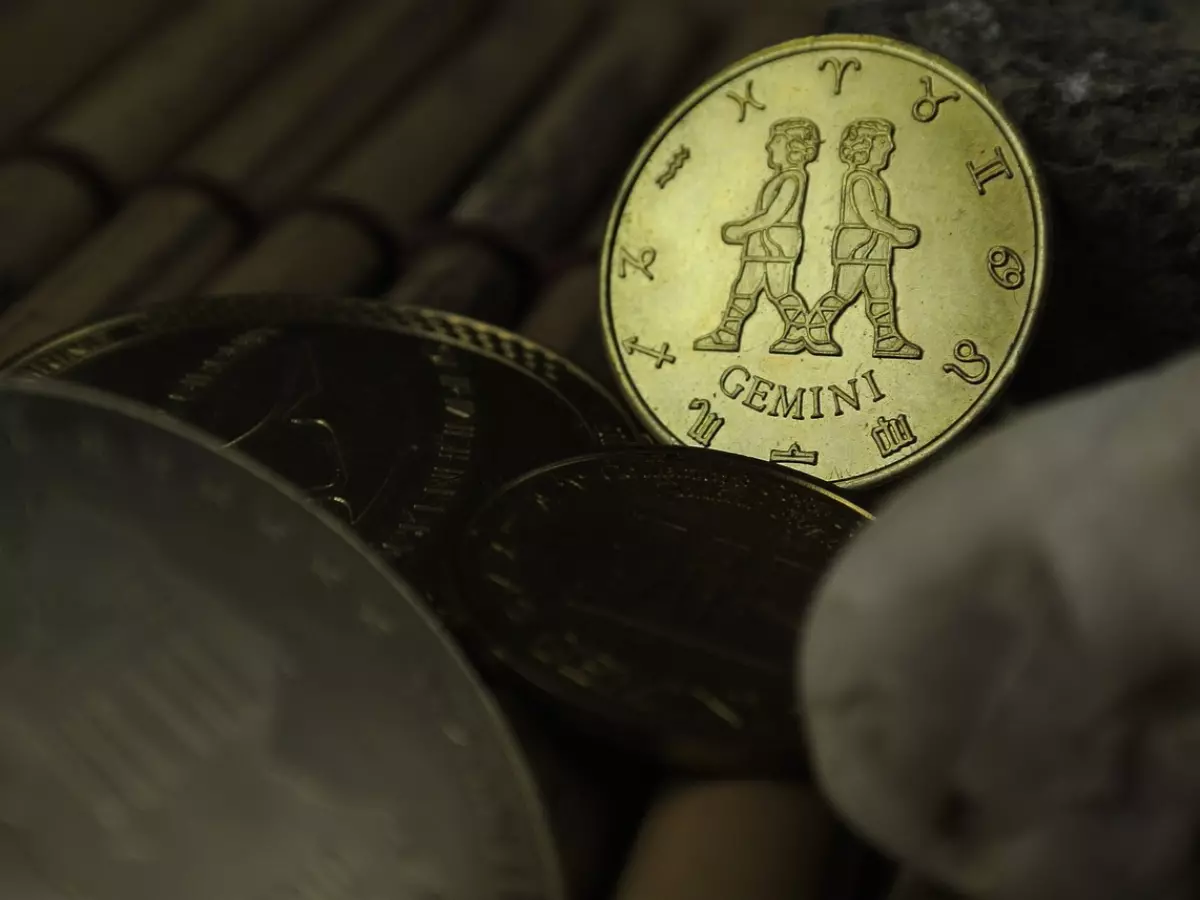 A close-up shot of a gold coin with the Gemini zodiac symbol. The coin is resting on a wooden surface and is surrounded by other coins.