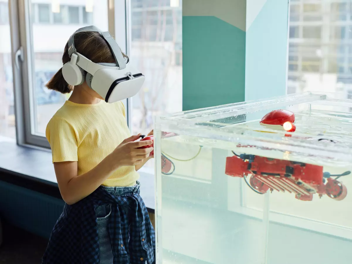 A person wearing a VR headset looks into a tank of water where a red robot is submerged. The robot is partially visible through the water.