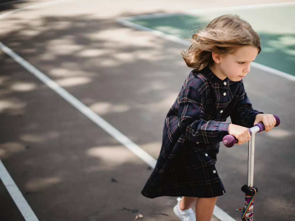 A little girl wearing a black dress with blue trim rides an electric scooter on a black paved track. She's moving to the right of the image.