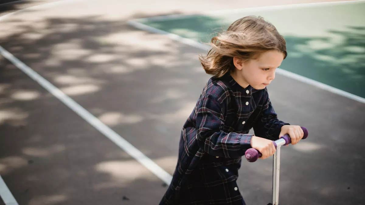 A little girl wearing a black dress with blue trim rides an electric scooter on a black paved track. She