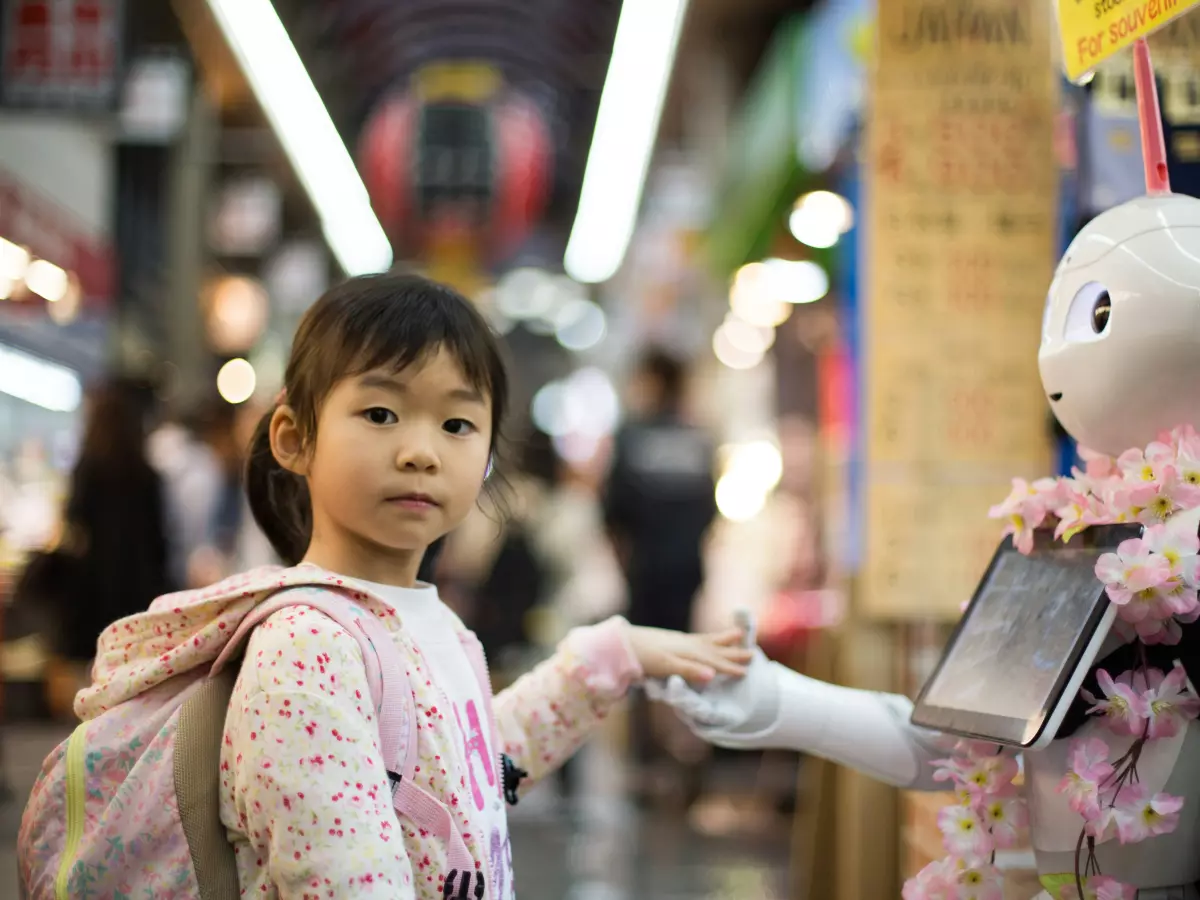 A young girl with a backpack stands facing a humanoid robot with a tablet. The robot is decorated with pink flowers and has a blue light around its eye.