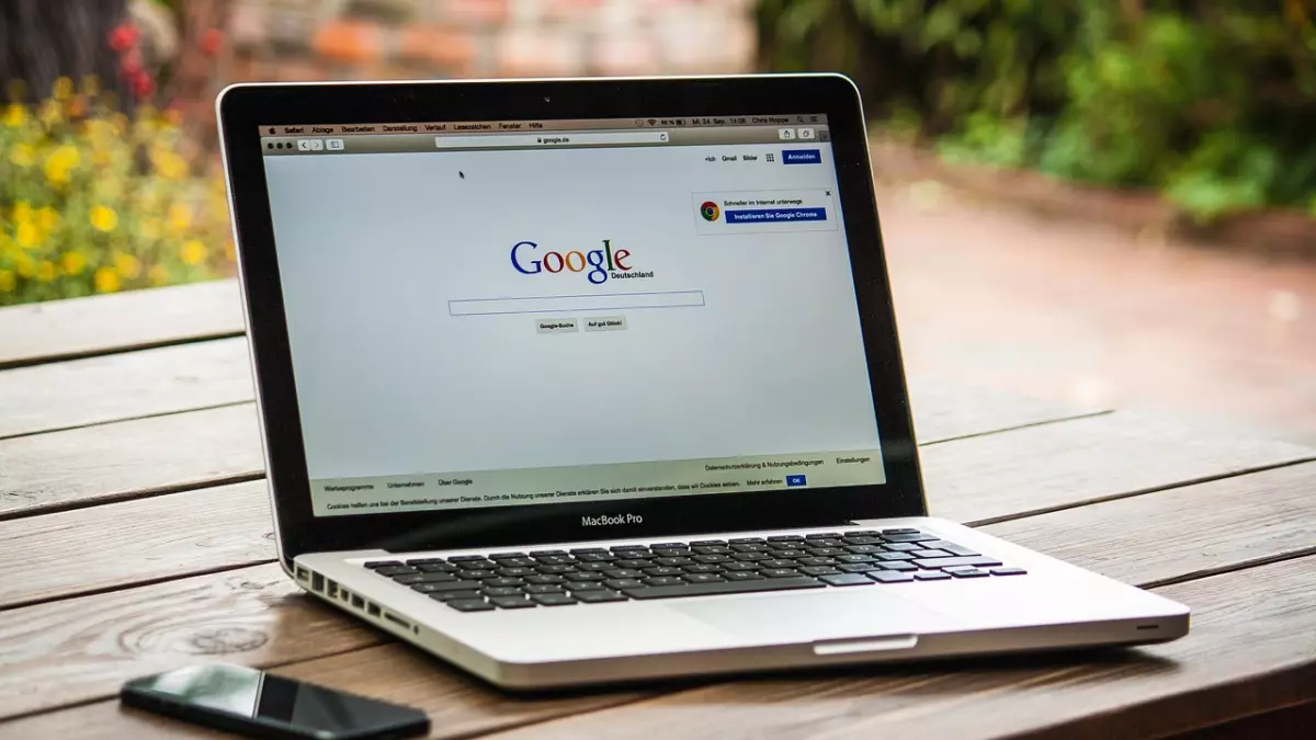 A silver Macbook Pro laptop is open on a wooden table. The laptop screen is showing a Google search page. A smartphone is on the table beside the laptop.