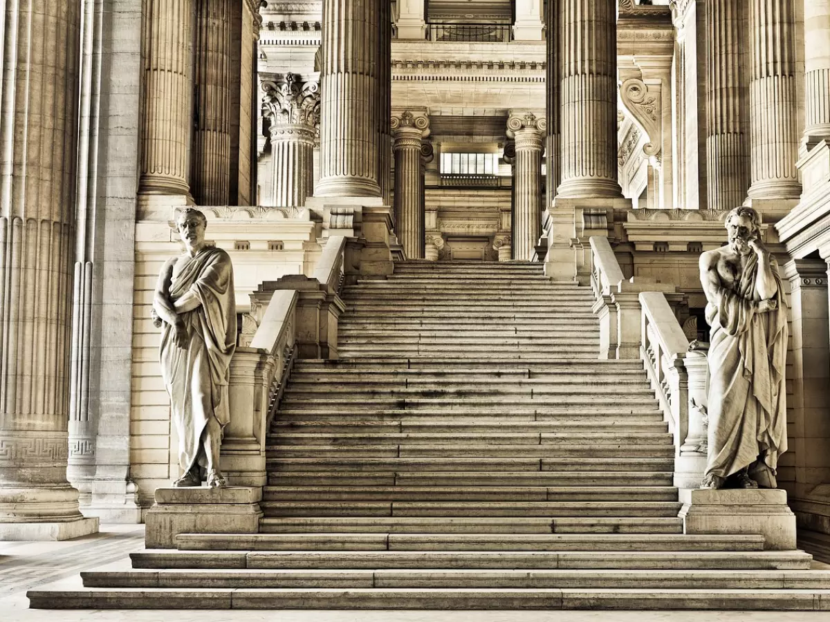 A grand staircase leading to a high ceiling with numerous columns. Two statues of men in togas stand at the bottom of the stairs. The image has a vintage filter applied to it, giving it a warm, sepia tone.