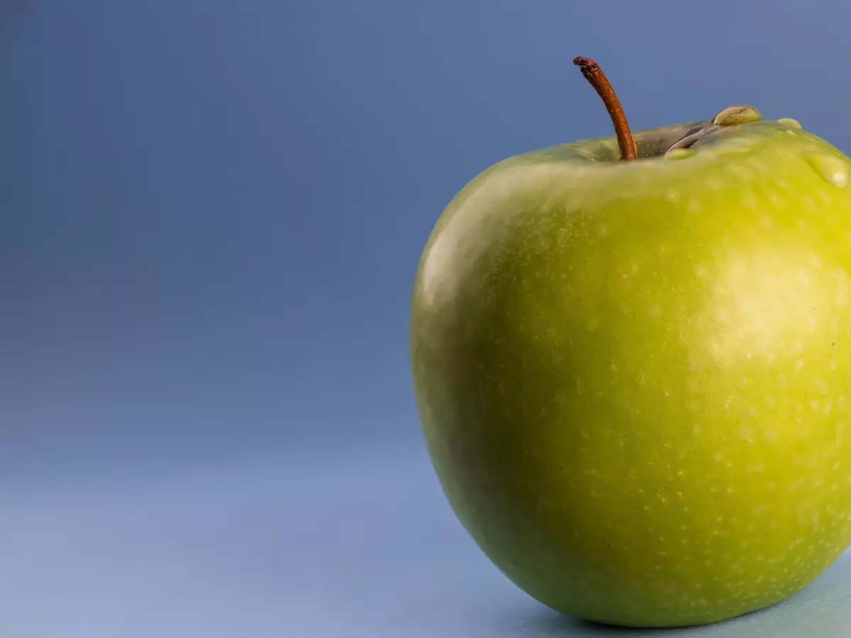 A close-up of a green apple with a single water drop on its surface. The apple is positioned against a soft blue background. 