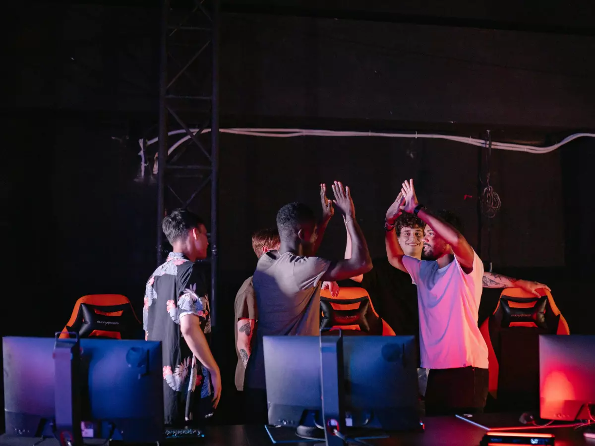 A group of young men, possibly gamers, are celebrating a victory while standing in front of a gaming station set up in a dark room with red lighting. The setting creates an immersive and exciting atmosphere.