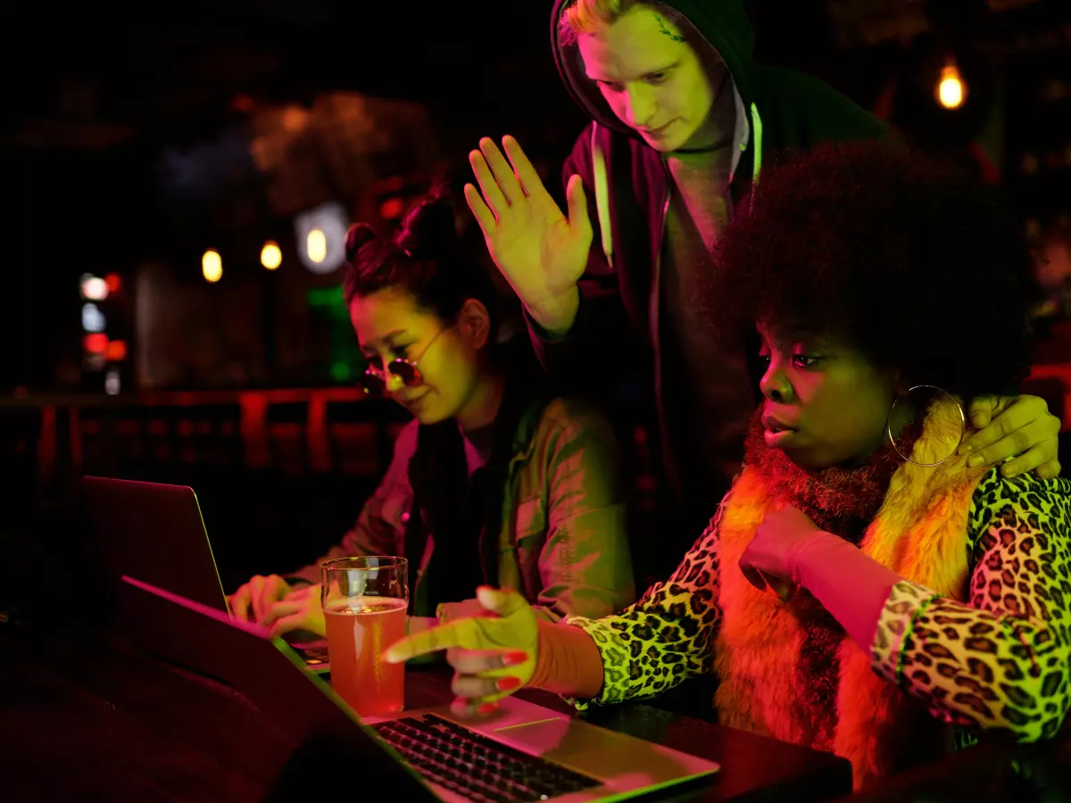 Three young women huddled around a laptop in a dimly lit bar setting. One woman gestures emphatically.