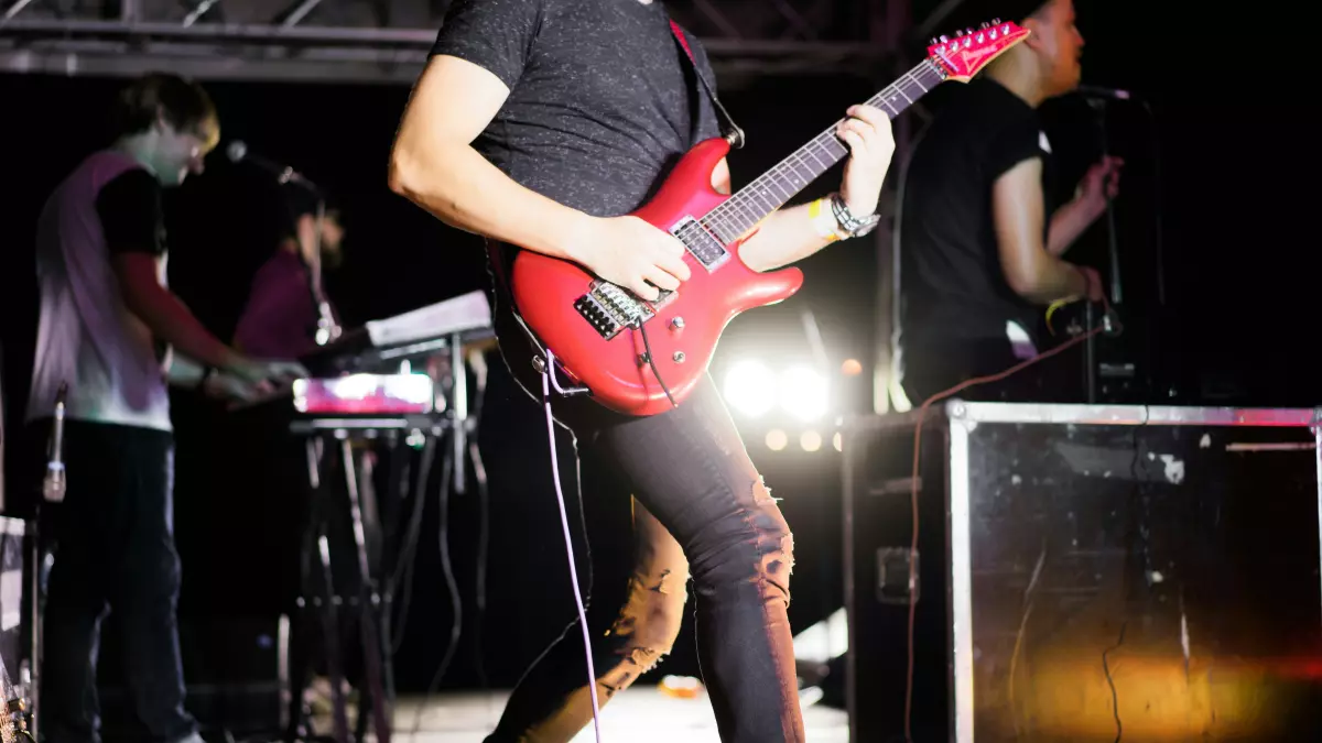 A band playing live music on a stage. The guitarist is in the foreground, and the drummer and keyboard player are in the background.