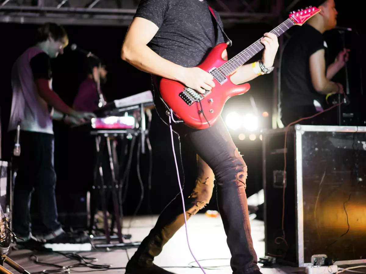 A band playing live music on a stage. The guitarist is in the foreground, and the drummer and keyboard player are in the background.