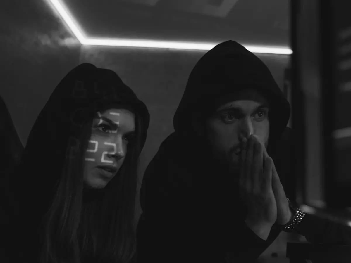 Two hooded figures sit in a dimly lit room, their faces obscured. The figures are in front of a computer screen with text displayed on it.