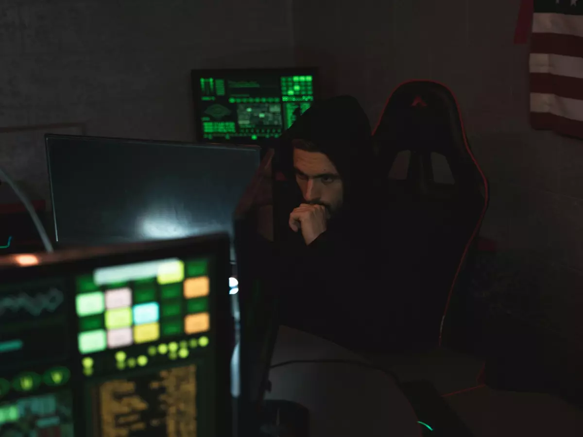 A hooded figure sits in front of multiple computer screens in a dark room, with an American flag hanging in the background. The figure's face is partially obscured, but they appear to be intensely focused on their work.