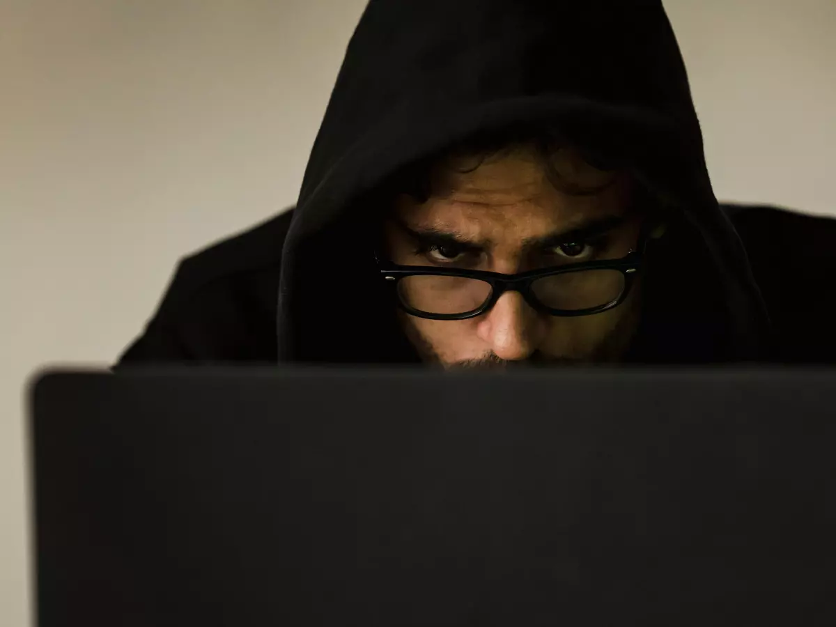 A person wearing a black hoodie and glasses is looking at a laptop. Their face is obscured by the hood, but their eyes are visible and focused on the screen. The background is blurry.