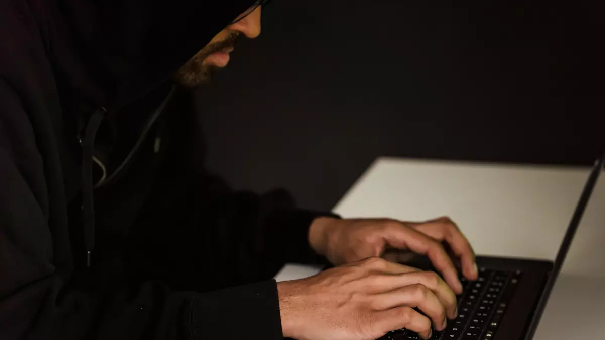 A person with their face hidden by a hood, typing on a laptop computer.