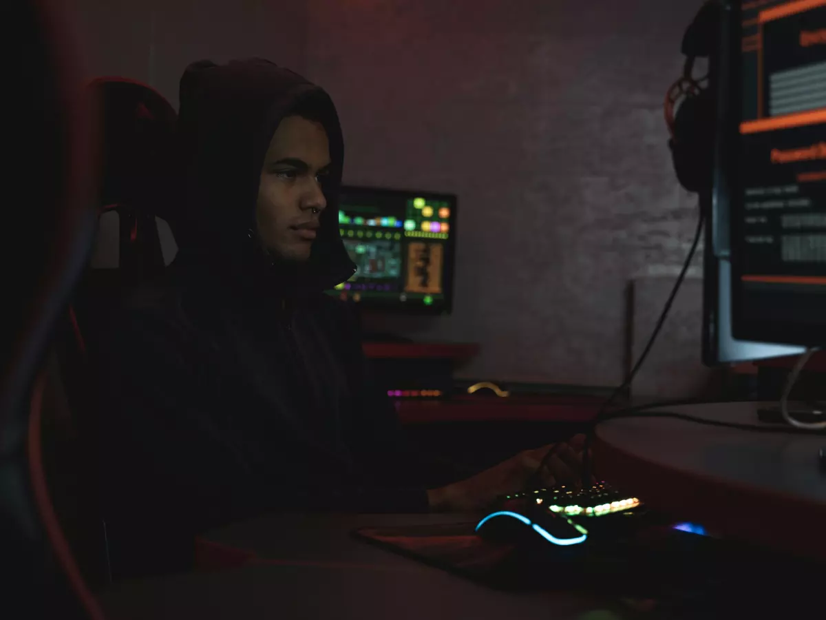 A hooded figure sits in front of a computer, typing intently, with a focus on the screen.