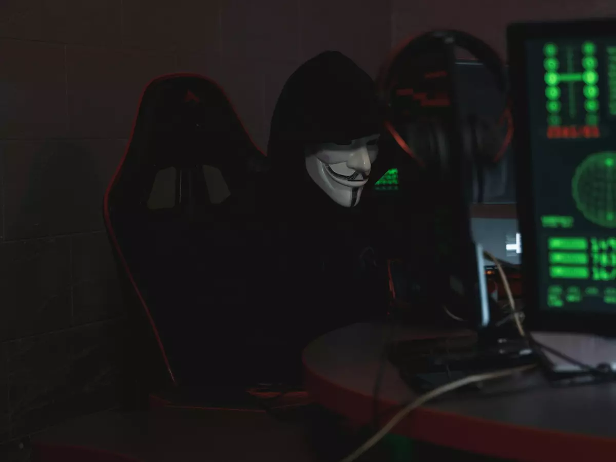 A hooded figure sits in front of a computer with a green screen, obscured by shadows, creating an air of mystery and danger.