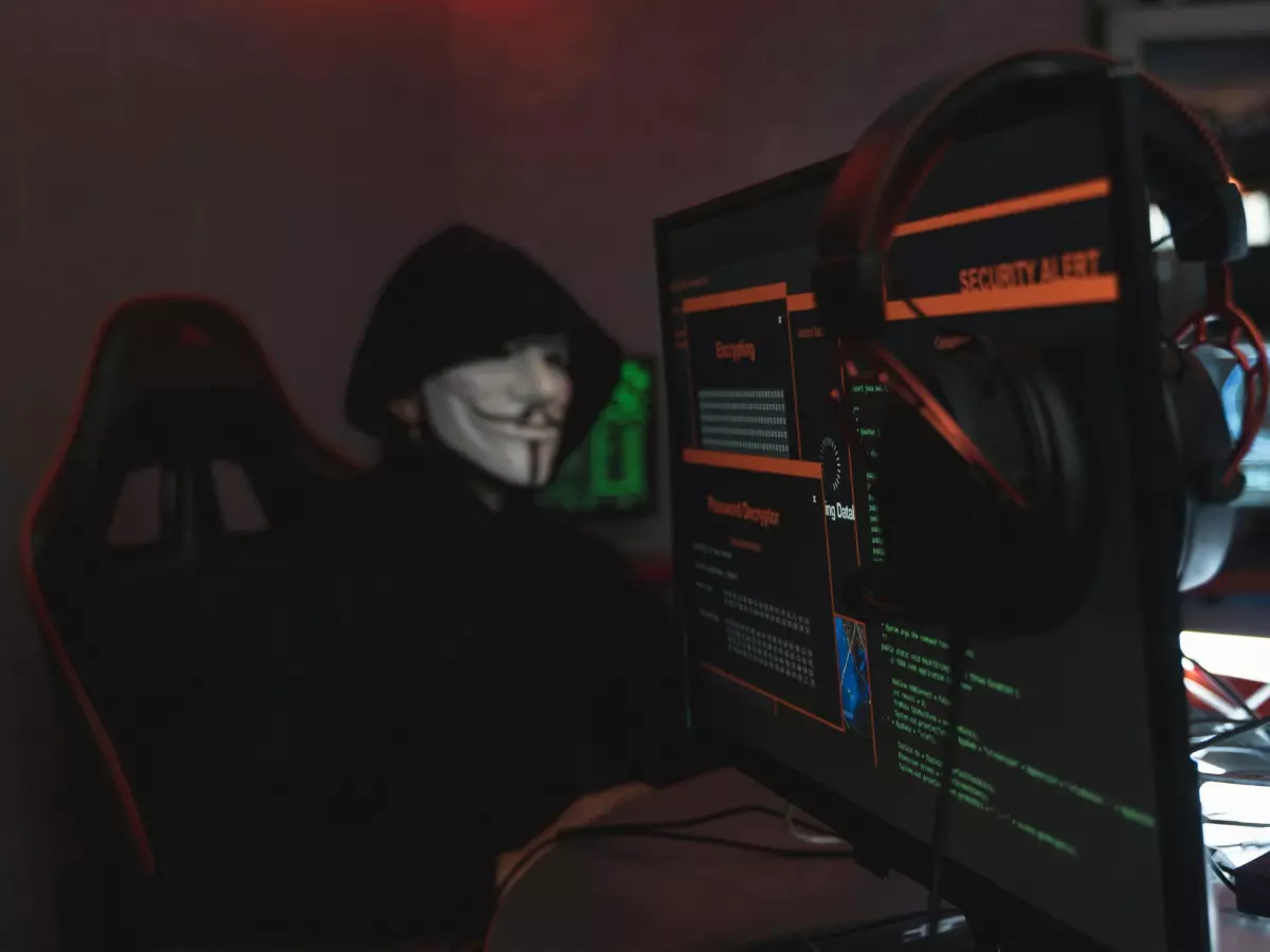 A hooded figure in a dark room, sitting in front of a computer screen with green and red code on it, looking sinister.