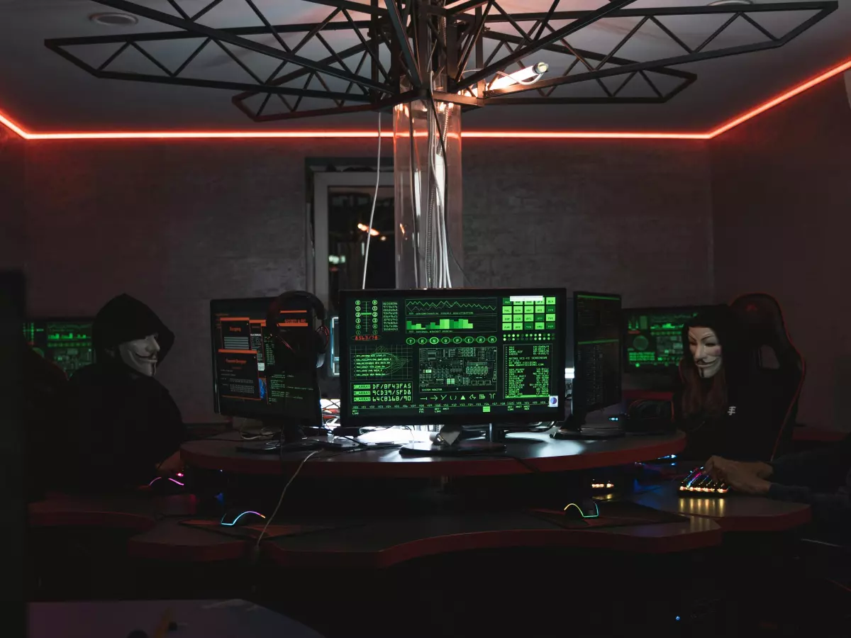 A group of hackers, wearing hoodies, are working in a dark room. They are surrounded by computer screens displaying code and data.