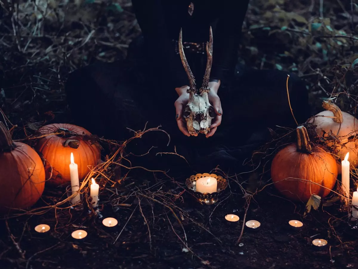 A woman in a black dress sits in a dark forest with a skull in her hands. She is surrounded by pumpkins and candles, which illuminate the scene and create a sense of mystery and magic. The image is evocative and dramatic, and it perfectly captures the dark and eerie atmosphere of the article.