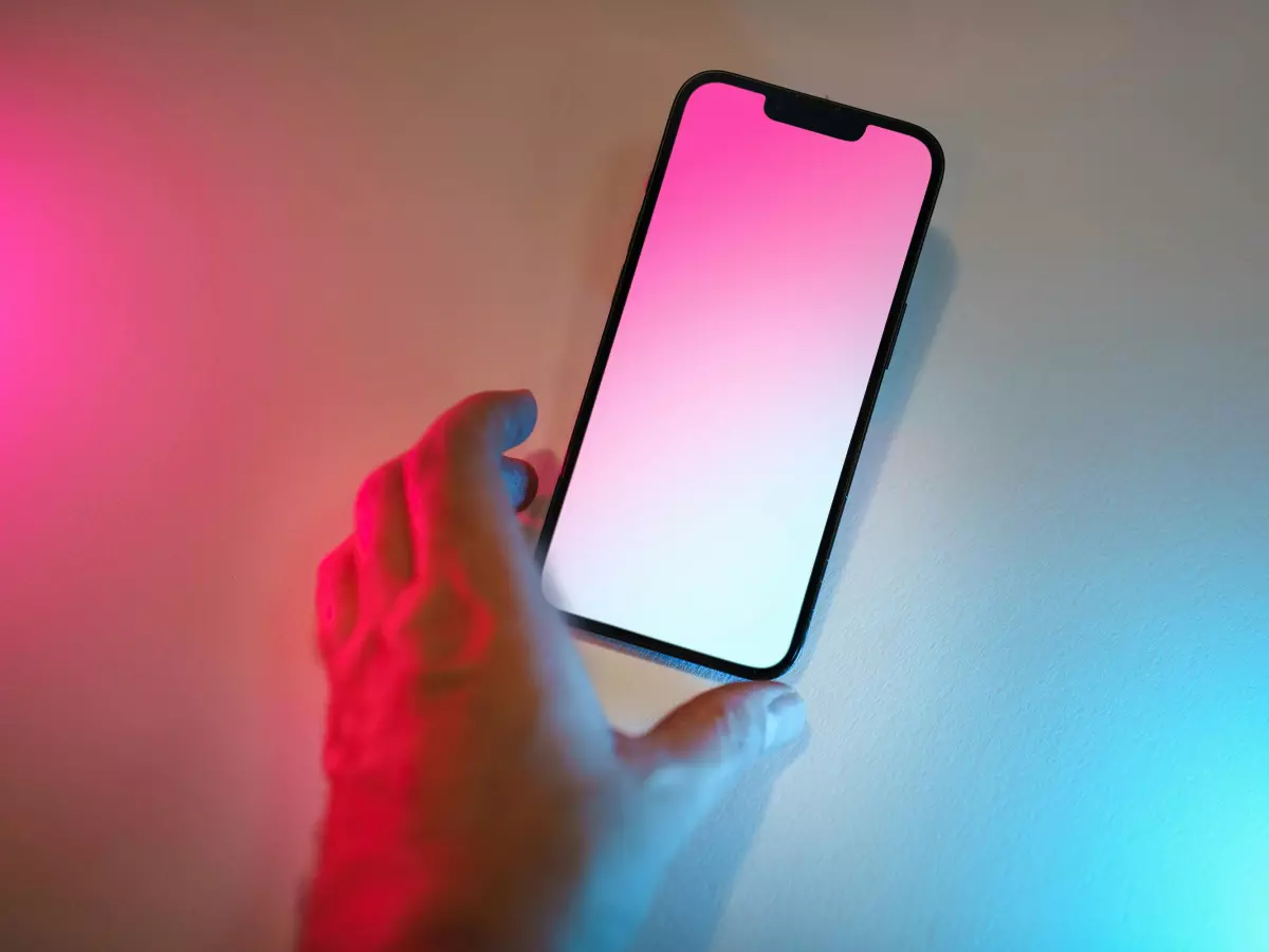 A hand holding a smartphone with a blank screen, lit by pink and blue lights.