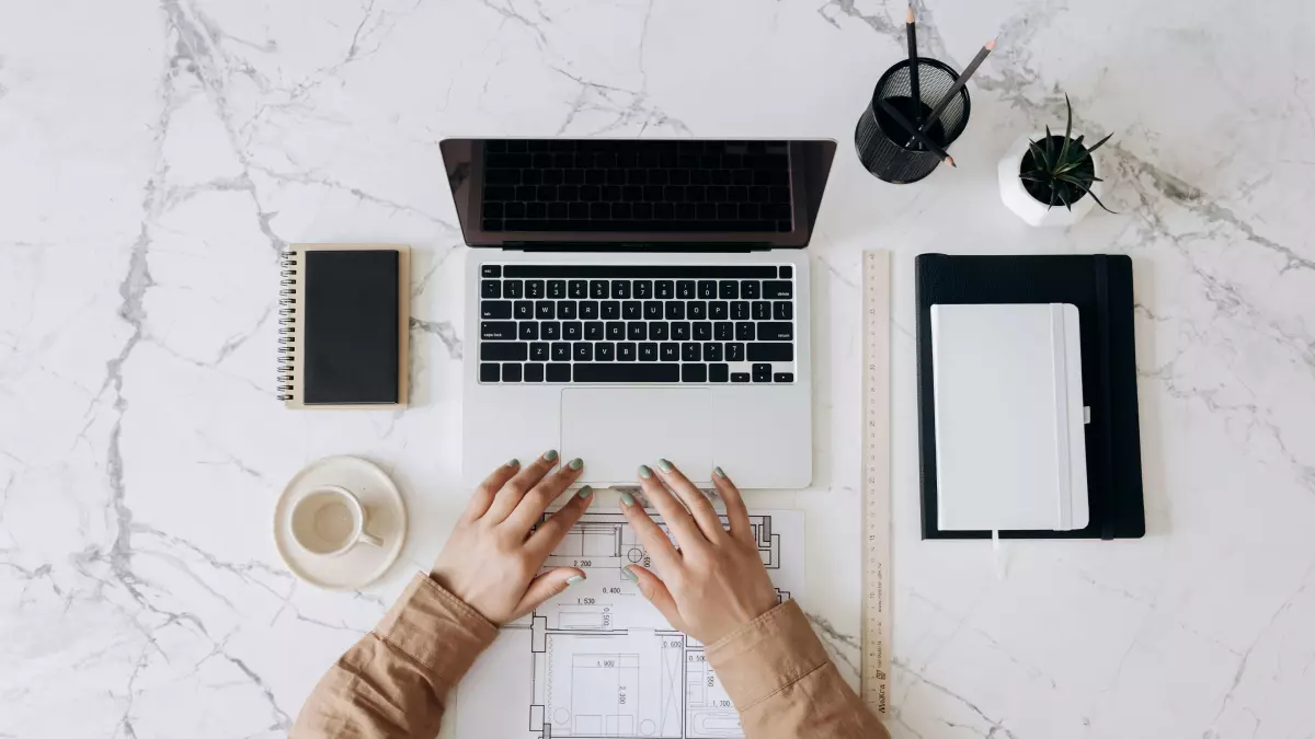 A person is sitting at a desk, using a laptop. They are wearing a light brown shirt and have their hands on the keyboard. The background is a white marble surface with a variety of office supplies.