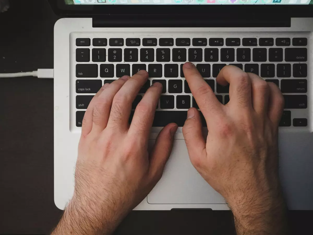 A close-up shot of a person's hands typing on a laptop keyboard. A white USB-C cable is connected to the laptop.