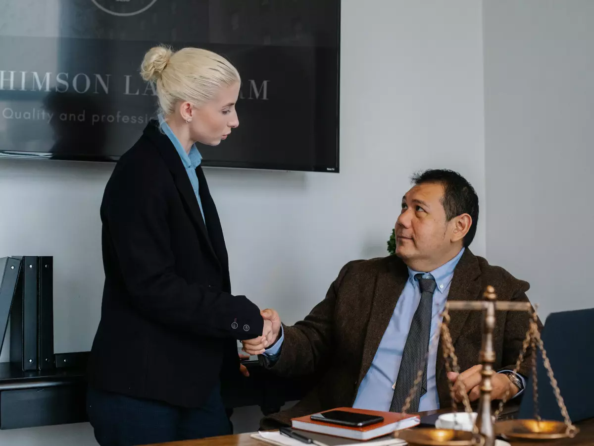 A woman in a business suit shakes hands with a man in a suit, who is sitting at a desk and holding a scale of justice.