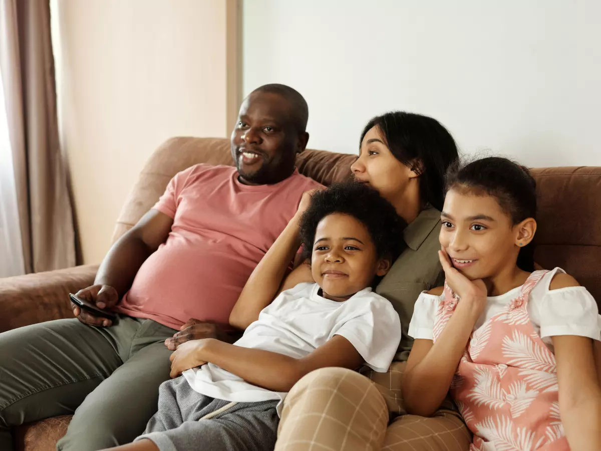 A family of four, an adult couple and two children, is sitting on a couch and watching TV. The family is smiling and enjoying their time together. The image is set in a living room.