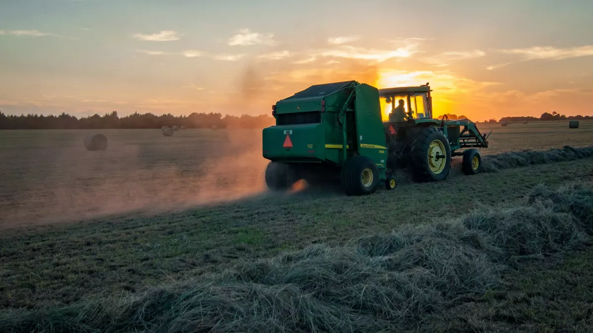 A man driving a green tractor through a field, baling hay. The sun is setting in the background.