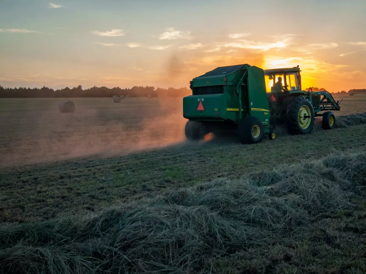 A man driving a green tractor through a field, baling hay. The sun is setting in the background.