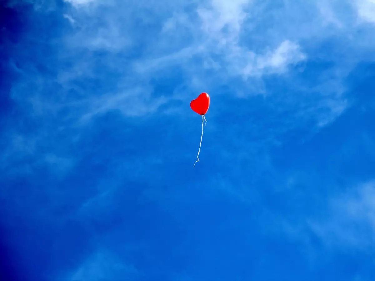 A red heart-shaped balloon floating in a blue sky with white clouds.